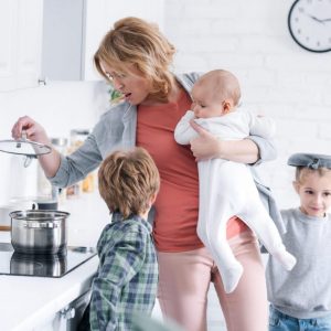 exhausted mother holding infant kid and cooking while naughty children playing in kitchen